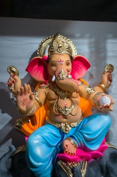 A colorful statue of a deity with an elephant head and multiple arms, adorned with gold jewelry. The figure is seated, with a decorative headdress and vibrant clothing in blue, pink, and orange hues. The hands of the statue hold various objects, and one hand is raised in a gesture of blessing.