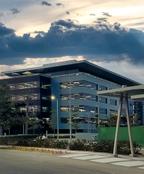 A modern office building with a geometric design is set against a dramatic sky filled with dark clouds. The structure features multiple floors with large windows, and the exterior is a mix of dark and light panels. In the foreground, there are trees and plants along with a crosswalk leading to the building.