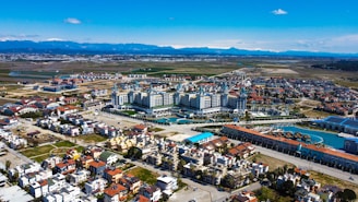 Aerial view of a complex resort situated amid a sprawling urban and suburban area. The resort has expansive buildings with ornate architecture, surrounded by pools and landscaped areas. The surrounding neighborhood consists of residential buildings with a mix of traditional and contemporary styles. Beyond the buildings, the landscape transitions into open fields and distant mountains under a clear blue sky.