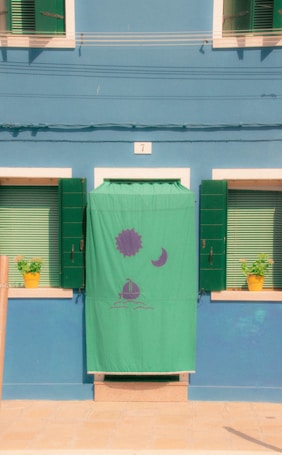 A brightly painted blue building facade features a doorway covered with a green cloth bearing simple images of a sun, a moon, and a sailboat. The green window shutters frame striped blinds on either side of the doorway, and potted plants in yellow pots add a touch of life to the scene.