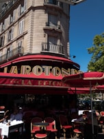 A classic Parisian street scene featuring an elegant stone building with wrought-iron balconies. The ground floor hosts a traditional café with a red and gold sign reading 'La Rotonde'. The outdoor seating consists of red wicker chairs and round tables, some occupied by patrons enjoying their drinks. A bright blue sky and lush green trees create a vibrant atmosphere.