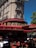 A classic Parisian street scene featuring an elegant stone building with wrought-iron balconies. The ground floor hosts a traditional café with a red and gold sign reading 'La Rotonde'. The outdoor seating consists of red wicker chairs and round tables, some occupied by patrons enjoying their drinks. A bright blue sky and lush green trees create a vibrant atmosphere.