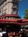 A classic Parisian street scene featuring an elegant stone building with wrought-iron balconies. The ground floor hosts a traditional café with a red and gold sign reading 'La Rotonde'. The outdoor seating consists of red wicker chairs and round tables, some occupied by patrons enjoying their drinks. A bright blue sky and lush green trees create a vibrant atmosphere.