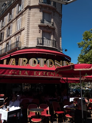 A classic Parisian street scene featuring an elegant stone building with wrought-iron balconies. The ground floor hosts a traditional caf&eacute; with a red and gold sign reading 'La Rotonde'. The outdoor seating consists of red wicker chairs and round tables, some occupied by patrons enjoying their drinks. A bright blue sky and lush green trees create a vibrant atmosphere.