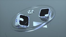 Two cube-shaped objects with black screens and white surfaces decorated with blue geometric patterns float within a large metallic ring. A compact keyboard and two game controllers accompany the cubes, all set against a minimalistic gray background.