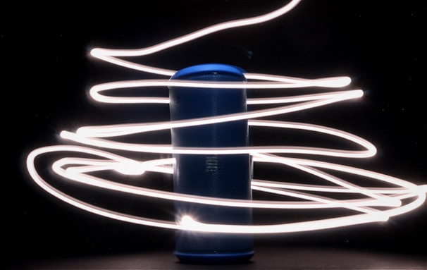 A cylindrical blue speaker stands in the center, surrounded by swirling trails of bright white light against a dark background. The light streaks create a dynamic and futuristic effect, as if wrapping around the speaker.