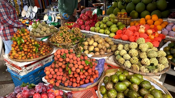 An outdoor market stall displays a variety of fresh fruits, arranged in baskets. The assortment includes lychees, dragon fruits, longans, sapodillas, pomelos, oranges, and other tropical fruits. The scene is vibrant, with shoppers visible in the background.