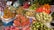 An outdoor market stall displays a variety of fresh fruits, arranged in baskets. The assortment includes lychees, dragon fruits, longans, sapodillas, pomelos, oranges, and other tropical fruits. The scene is vibrant, with shoppers visible in the background.
