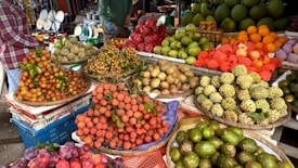 An outdoor market stall displays a variety of fresh fruits, arranged in baskets. The assortment includes lychees, dragon fruits, longans, sapodillas, pomelos, oranges, and other tropical fruits. The scene is vibrant, with shoppers visible in the background.
