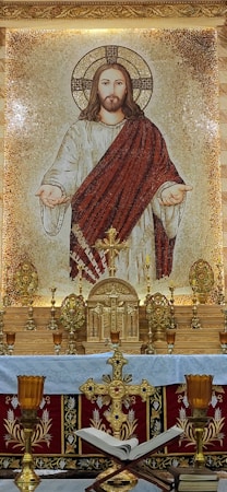 A mosaic depicting a religious figure with outstretched arms, wearing a white robe with a red drape, and a halo around the head. The figure is surrounded by ornate golden decorations, including a cross, candelabras, and floral designs. In the foreground, there is an altar adorned with liturgical items such as crosses, candles, and an open book.