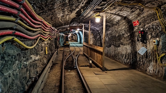 A dimly lit tunnel is reinforced with stone walls and has railway tracks running through its center. Multiple cables are attached along the left wall, and a light fixture hangs from the ceiling. A wooden barrier divides the path, and equipment like an emergency phone and fire extinguisher sign are visible on the right wall.