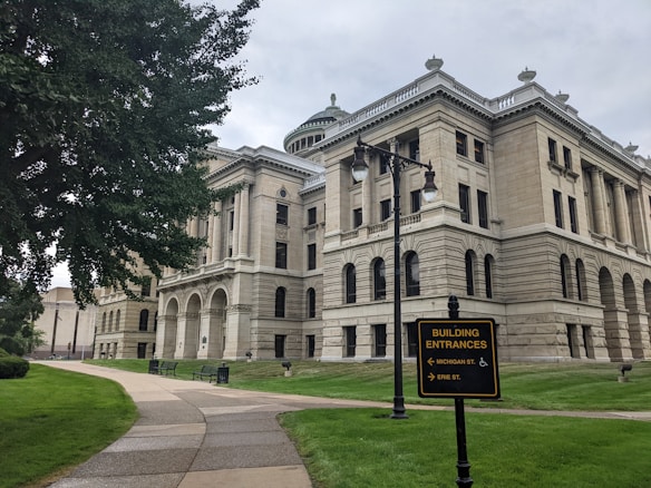 A large, historic-looking building with classical architecture featuring columns and arched windows. A well-maintained lawn surrounds the building, and a tree stands on the left side. A pathway with benches leads towards the building. There's a sign in the foreground indicating building entrances with directions to Michigan St. and Erie St.
