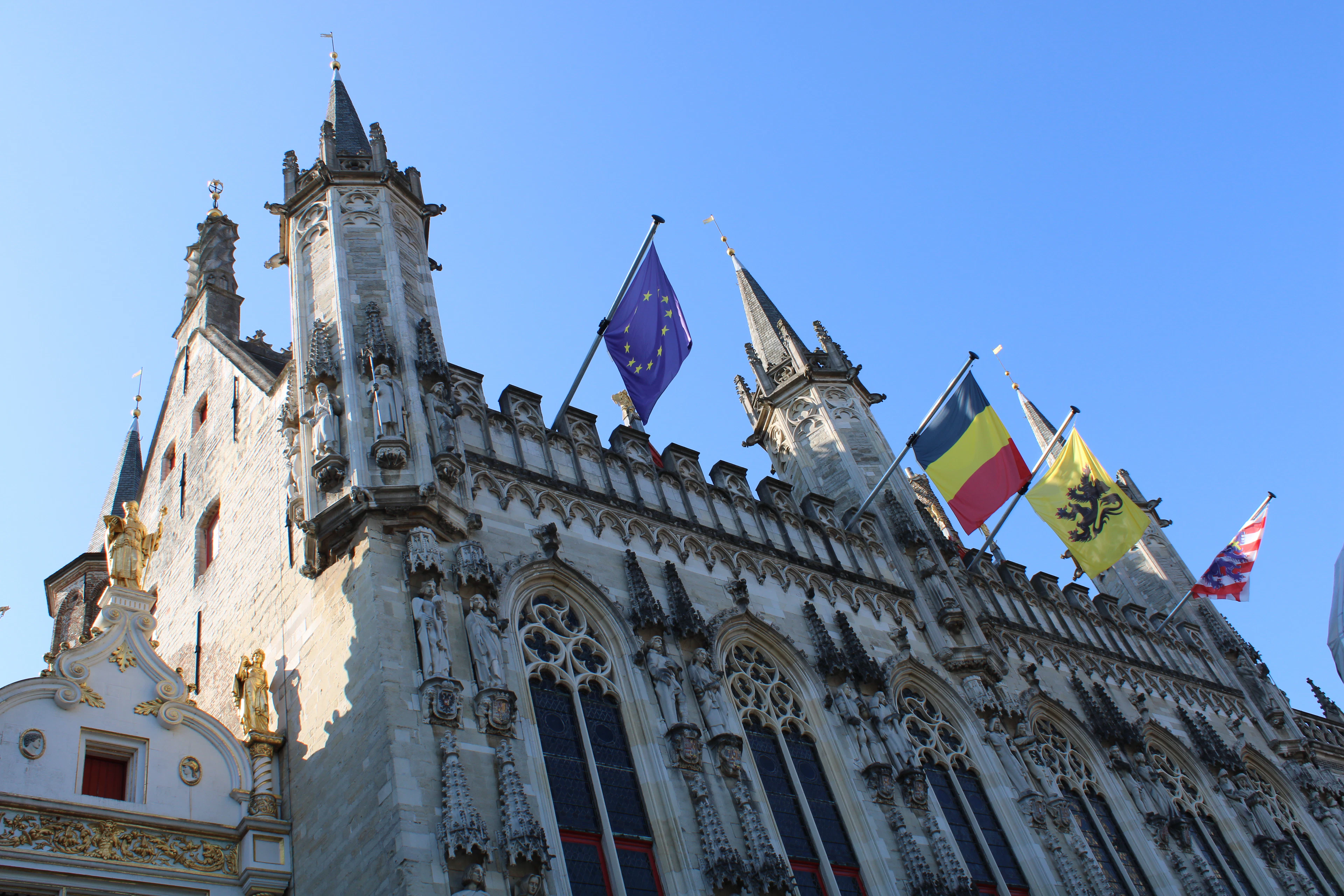 Ornate Gothic spires and flags atop a historic building on Bruges' Burg square against a clear blue sky.