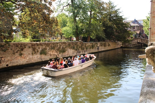 A group of people is enjoying a scenic boat ride along a canal surrounded by lush greenery and old brick walls. The waterway is calm, reflecting the sunlight and the canopy of trees overhead. A stone bridge is visible in the background, adding to the picturesque setting.