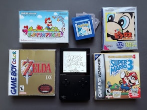 A collection of vintage video game cartridges and cases for different Nintendo systems, including Game Boy, Game Boy Color, and Game Boy Advance. Visible games are 'Super Mario Advance', 'The Legend of Zelda: Link's Awakening DX', 'Kirby's Dream Land', 'Super Mario Advance 4: Super Mario Bros. 3', and a 'Pokemon' cartridge. The layout suggests a nostalgic gaming theme.
