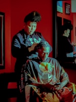A person with braided hair styles another individual's hair in a salon. The setting features a rich red color palette with a mirror reflecting the scene. Both individuals are focused, with the seated person using a phone.