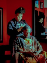 A person with braided hair styles another individual's hair in a salon. The setting features a rich red color palette with a mirror reflecting the scene. Both individuals are focused, with the seated person using a phone.