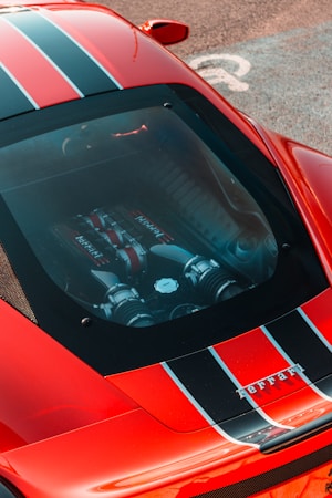 A red sports car with black and white racing stripes on the body is shown with a focus on the engine visible through a transparent hood. The car is parked on a textured surface, and a part of a parking space marking is visible.