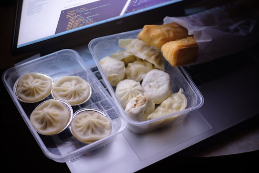 A variety of dumplings and fried bread are arranged in transparent plastic containers on top of a laptop keyboard. The image is dimly lit, suggesting a relaxed or casual setting. Some dumplings are placed in foil cups, while others are loosely packed. The laptop screen displays some coding text or script, adding a tech-savvy ambiance.