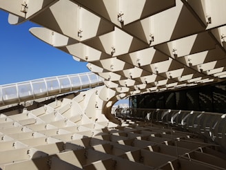 The image features a large modern architectural structure composed of numerous geometric patterns and panels. The structure's design presents a complex interplay of light and shadow due to its open, partially transparent surfaces. The bright blue sky can be seen in the background, contrasting with the beige tones of the architecture.