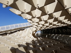 The image features a large modern architectural structure composed of numerous geometric patterns and panels. The structure's design presents a complex interplay of light and shadow due to its open, partially transparent surfaces. The bright blue sky can be seen in the background, contrasting with the beige tones of the architecture.