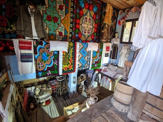 A room filled with traditional textiles, embroidered clothing, and wooden artifacts. The walls are adorned with colorful tapestries featuring intricate floral and geometric patterns. Various articles of clothing, including embroidered shirts and jackets, are displayed on hangers. Wooden barrels and household items like pottery and woven baskets are scattered around the room, adding to the rustic atmosphere.