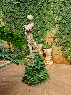 A classical stone statue of a woman holding a small object stands in a lush garden setting. The statue is partially covered with creeping ivy. The backdrop features a wall adorned with dense green foliage, and there are two decorative pots with ferns beside the statue.