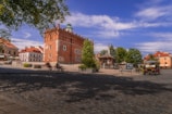 A charming town square with a prominent historical red-brick building featuring a green dome. The square is paved with cobblestones and surrounded by trees and smaller buildings with red roof tiles. There are benches, flower pots, and a few people enjoying the open space. A decorative rotunda stands in the center, and a small vehicle is parked nearby.