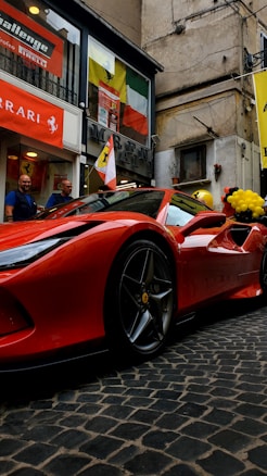 A sleek red sports car is parked on a cobblestone street next to a building adorned with various Ferrari flags and banners. Two people stand nearby, and there are black and yellow balloons in the background.