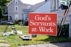 A red sign with white text reads 'God's Servants at Work', placed on a grassy area in front of a residential house. The background contains a ladder, some paint supplies, and garden tools leaning against a structure, suggesting ongoing house maintenance or renovation.