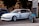 A modified white hatchback car is parked next to a customized blue motorcycle. The car has sporty features, including branded decals and the number 13 on the side, along with text that reads 'No Good Racing.' Nearby, a modern parking sign provides directional information. The background features trees and institutional buildings, possibly indicating an urban environment.