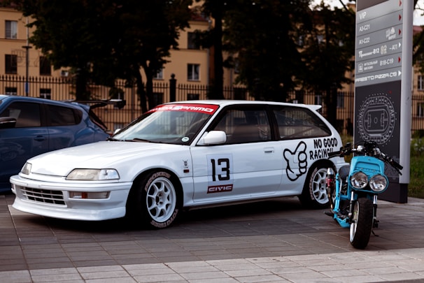 A modified white hatchback car is parked next to a customized blue motorcycle. The car has sporty features, including branded decals and the number 13 on the side, along with text that reads 'No Good Racing.' Nearby, a modern parking sign provides directional information. The background features trees and institutional buildings, possibly indicating an urban environment.
