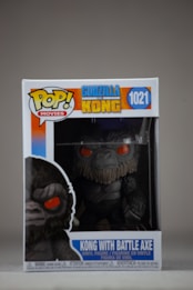 A boxed Funko Pop! vinyl figure of Kong holding a battle axe, from the movie Godzilla vs. Kong. The packaging displays vibrant colors and features a large image of the character with red eyes. The figure itself is visible through clear plastic within the box.