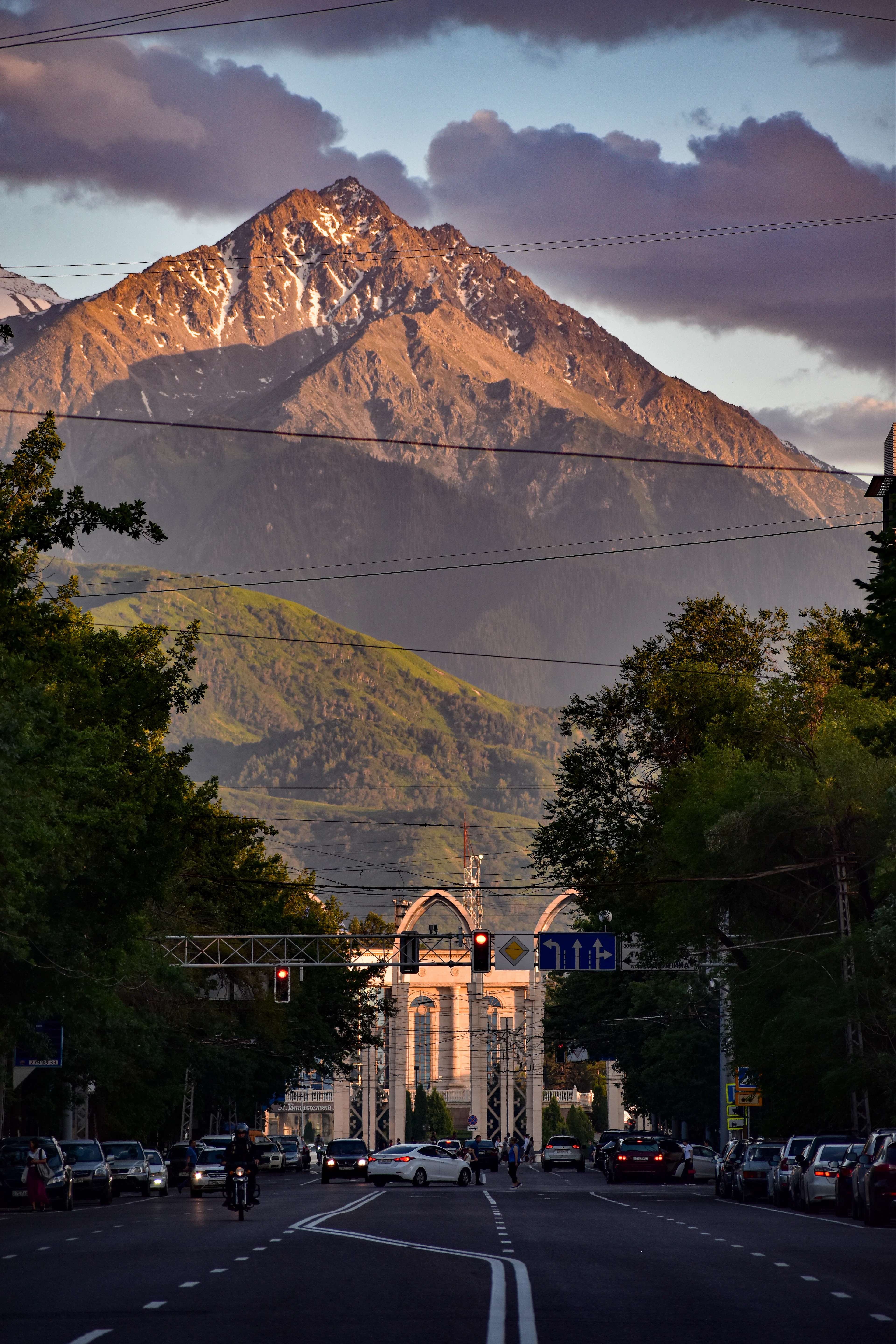 A sunset-lit mountain peak looms over an Almaty avenue with a stone gateway and evening traffic.