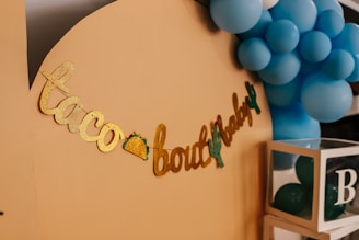 A decorative display featuring gold glitter lettering spelling out 'taco bout a baby' with small taco and cactus images integrated into the design. Blue balloons form a cluster in the background, providing a festive atmosphere. A transparent box filled with more balloons, labeled with the letter 'B', sits nearby.
