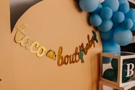 A decorative display featuring gold glitter lettering spelling out 'taco bout a baby' with small taco and cactus images integrated into the design. Blue balloons form a cluster in the background, providing a festive atmosphere. A transparent box filled with more balloons, labeled with the letter 'B', sits nearby.