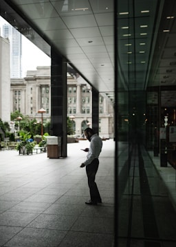 A man stands in a spacious, modern architectural setting, intently looking at his smartphone. The location is outdoors but sheltered, with a large glass facade reflecting part of the scene. In the background, trees, benches, and historic buildings are visible.