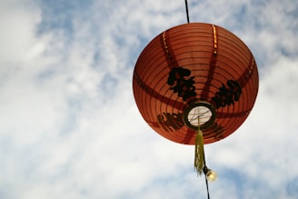 A bright red Chinese lantern with black calligraphy hanging against a partly cloudy sky. The lantern features decorative elements and a small tassel at the bottom, with a light bulb visible below it.