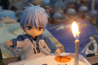 A cute anime-style figurine with blue hair and striking eyes wearing a blue and white sports jersey numbered 15. It is positioned next to a lit birthday candle on a cake decorated with a basketball symbol. The background includes several similar figurines and blurred objects, adding depth to the scene.