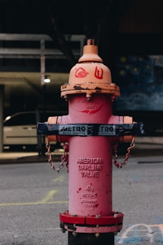 A pink fire hydrant stands on a city street with a dark background. The hydrant is wrapped with a metal chain and displays text that reads 'American Darling Valve'. There are painted signs above the text, possibly graffiti. The ground shows some faded markings, and a vehicle is partially visible in the background.