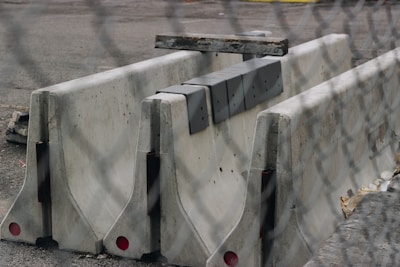 Several large concrete barriers are placed closely together on a paved surface. There are some metal brackets attached to the barriers, and the scene is viewed through a chain-link fence, which creates a blurred effect in the foreground.