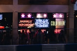 A bustling nightlife scene with neon signs lighting up a storefront. The signs display phrases like 'Nashville Hot Chicken,' 'Hello Good Times,' and 'Cold Beer.' A group of people is gathered outside, likely enjoying a night out on the town.