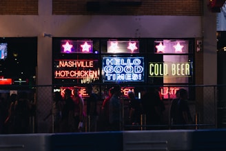 A bustling nightlife scene with neon signs lighting up a storefront. The signs display phrases like 'Nashville Hot Chicken,' 'Hello Good Times,' and 'Cold Beer.' A group of people is gathered outside, likely enjoying a night out on the town.