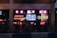 A bustling nightlife scene with neon signs lighting up a storefront. The signs display phrases like 'Nashville Hot Chicken,' 'Hello Good Times,' and 'Cold Beer.' A group of people is gathered outside, likely enjoying a night out on the town.