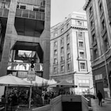 An urban scene featuring tall buildings with classical architectural elements. A pedestrian walkway is visible with outdoor seating under umbrellas. There is a sign in Spanish and a small cafe booth.