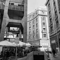 An urban scene featuring tall buildings with classical architectural elements. A pedestrian walkway is visible with outdoor seating under umbrellas. There is a sign in Spanish and a small cafe booth.