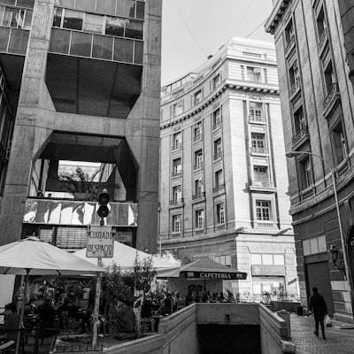 An urban scene featuring tall buildings with classical architectural elements. A pedestrian walkway is visible with outdoor seating under umbrellas. There is a sign in Spanish and a small cafe booth.