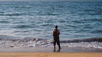 A person stands on a sandy beach, holding a fishing rod, facing the vast body of water with gentle waves approaching the shore. The water appears deep blue, blending into the horizon.