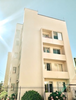 A multi-story residential building with a light cream facade under a clear blue sky. Sunlight is streaming from the top left corner, illuminating the building walls. Surrounding the building is a black metal fence with lush green bushes and pink flowers.