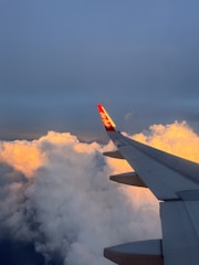 An airplane wing with a red AirAsia logo extends over a vast expanse of fluffy white clouds. The clouds are illuminated with a warm golden hue by the sunlight, contrasting with the clear blue sky above.